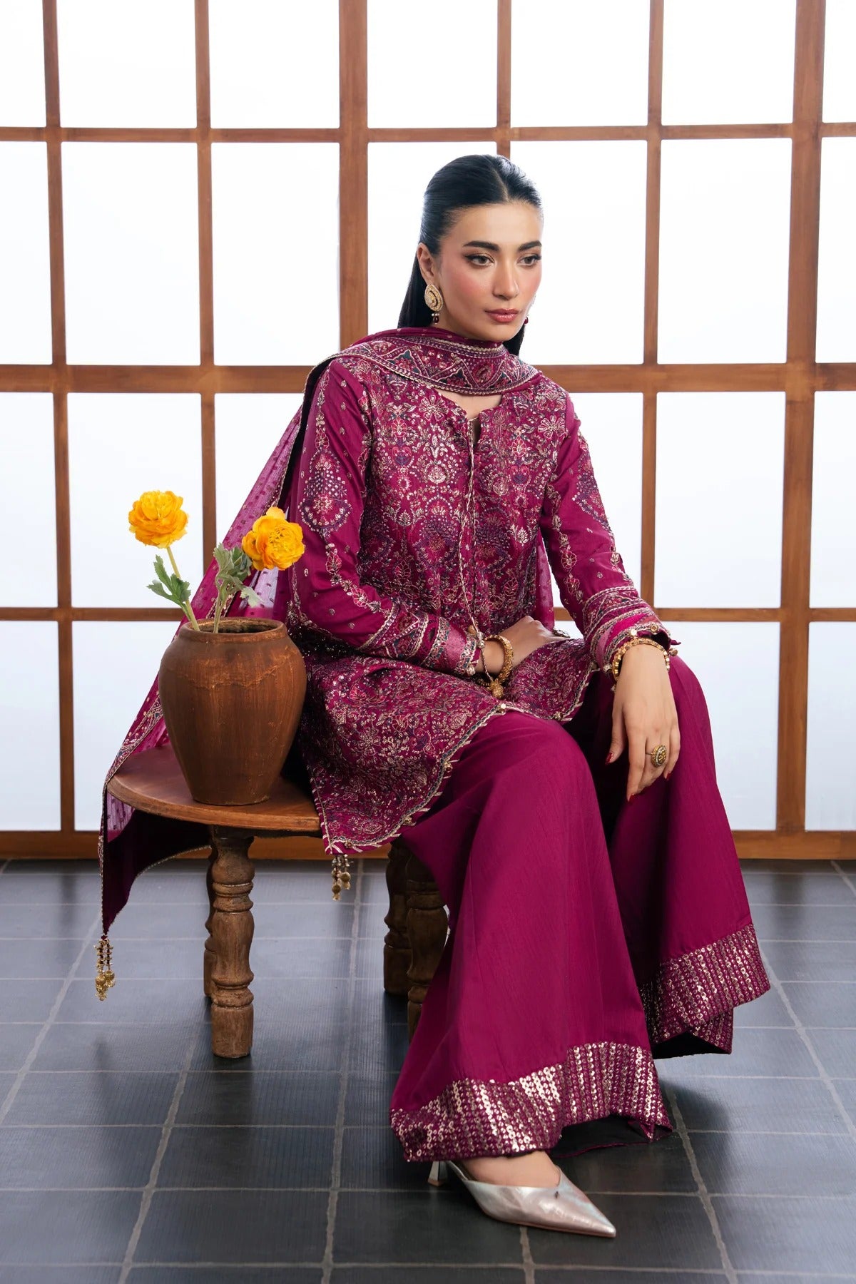 A woman models a traditional three-piece salwar kameez suit in a deep magenta-purple color with intricate gold embroidery, seated on a wooden stool in front of a wooden screen.