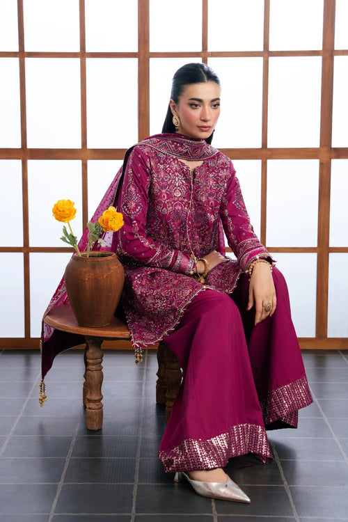A woman models a traditional three-piece salwar kameez suit in a deep magenta-purple color with intricate gold embroidery, seated on a wooden stool in front of a wooden screen.
