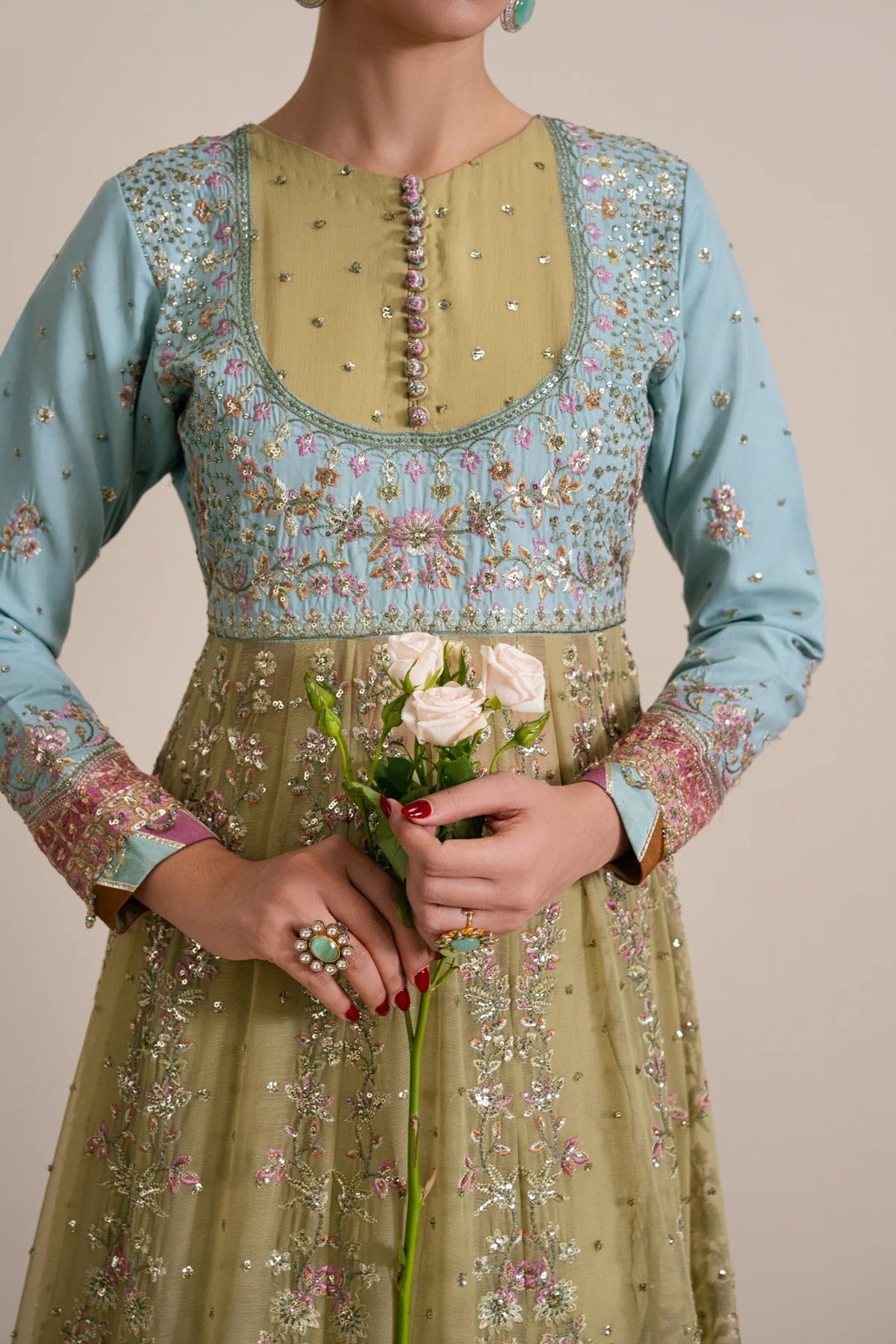 A woman wearing a light blue and olive green heavily embroidered traditional dress, holding a small bouquet of white roses against a plain beige background.