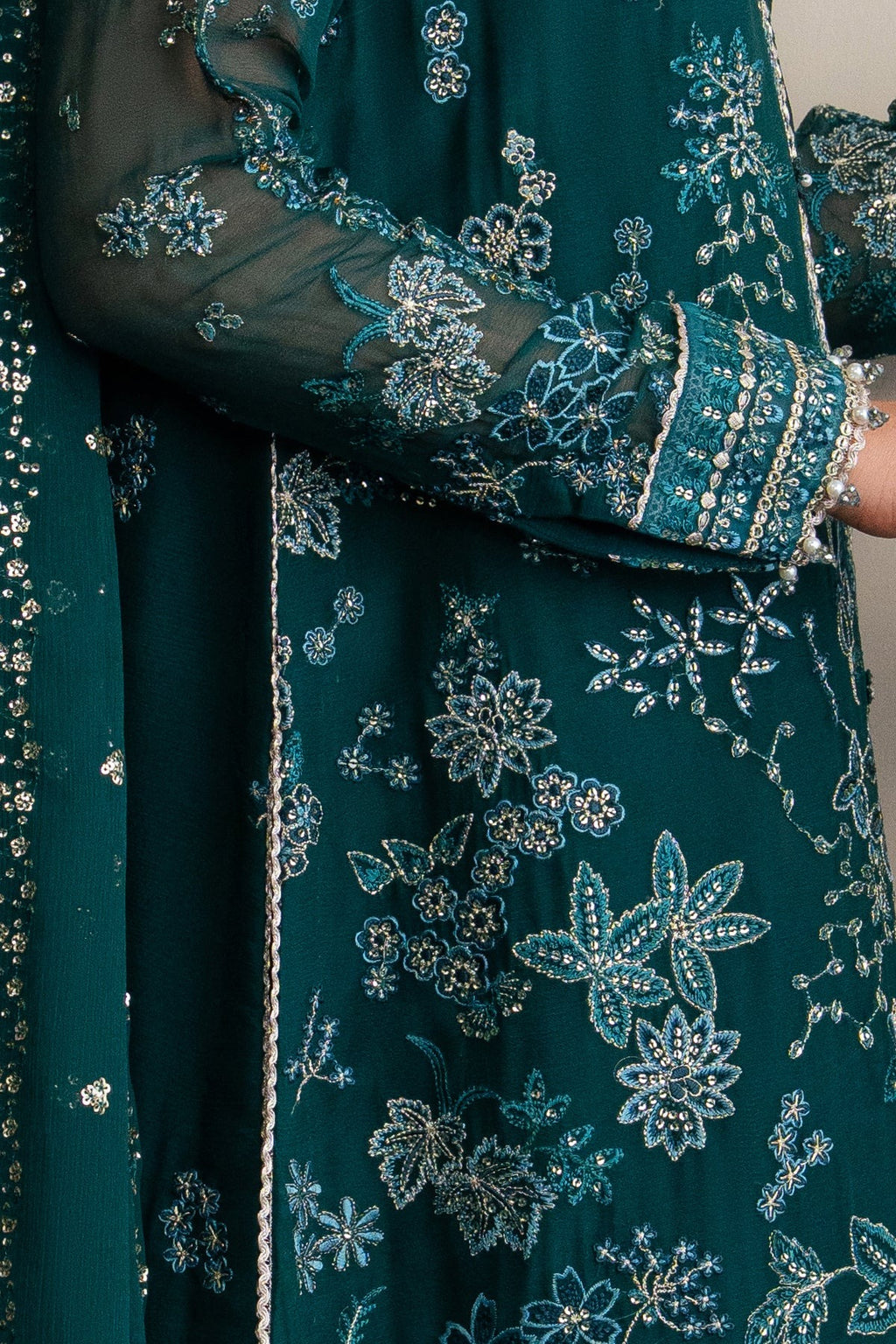 Close-up of a dark teal green traditional embroidered dress with intricate silver and blue floral embroidery on sheer sleeves and a matching dupatta.
