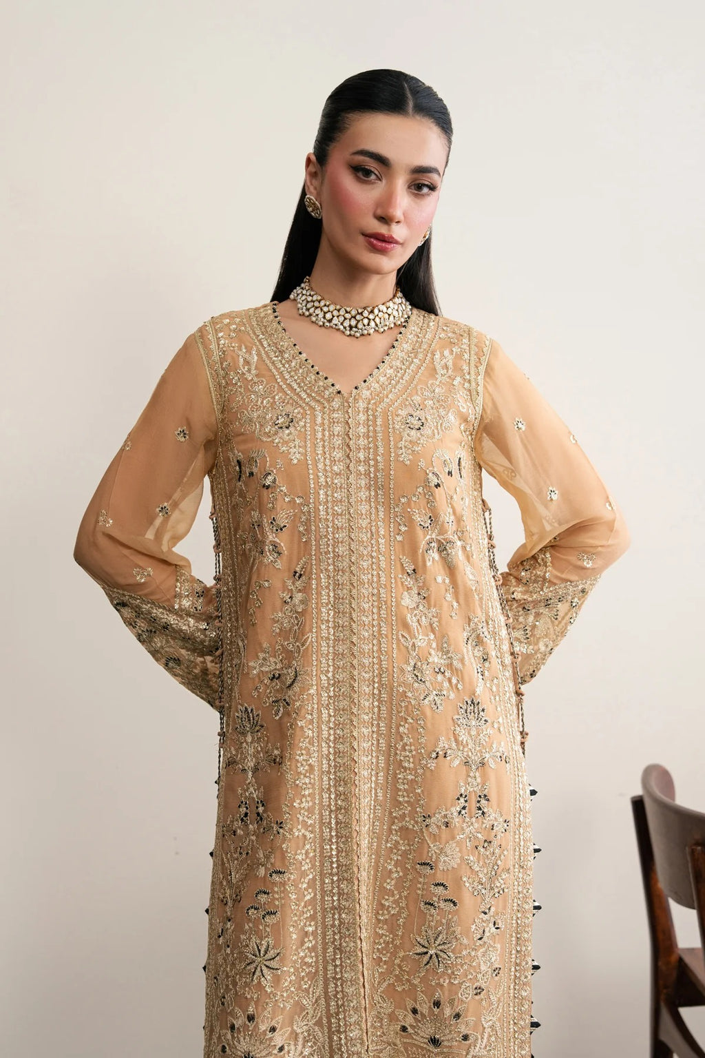 A woman modeling a beige embroidered traditional long kurta with sheer sleeves, intricate metallic embroidery, and beadwork, paired with a matching beaded choker necklace against a plain light background.