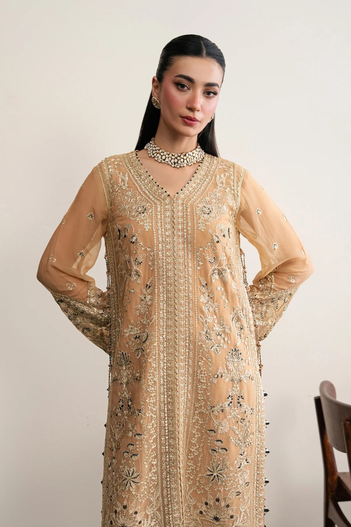 A woman modeling a beige embroidered traditional long kurta with sheer sleeves, intricate metallic embroidery, and beadwork, paired with a matching beaded choker necklace against a plain light background.
