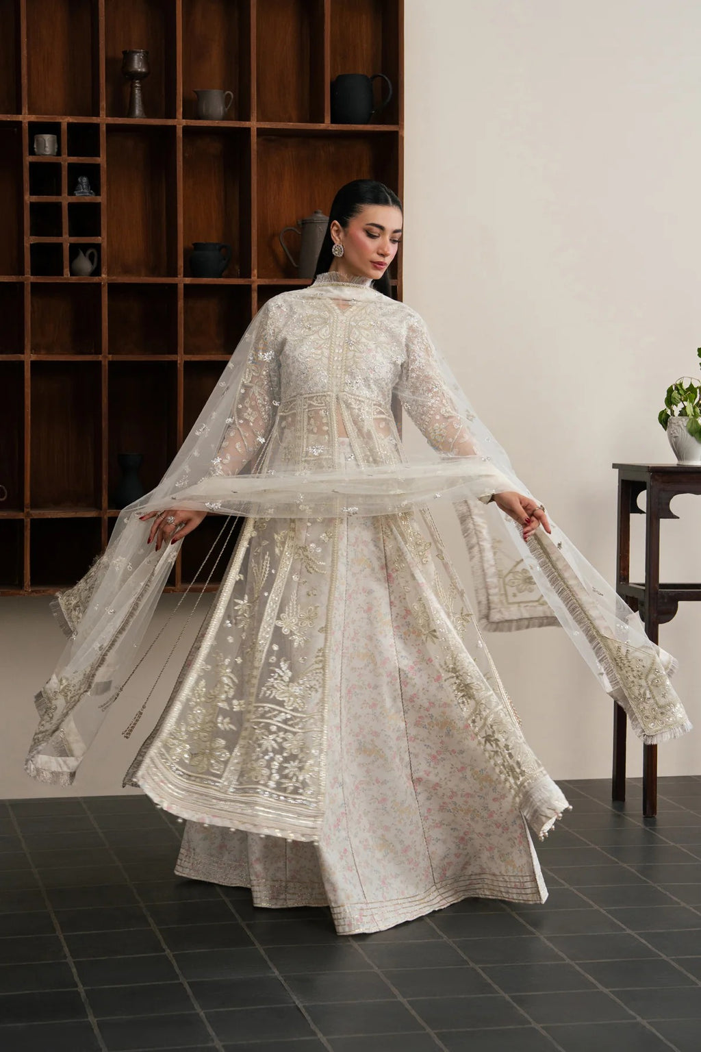 A woman wearing a traditional white embroidered Anarkali gown with a matching sheer dupatta, posing in a room with a wooden bookshelf in the background.
