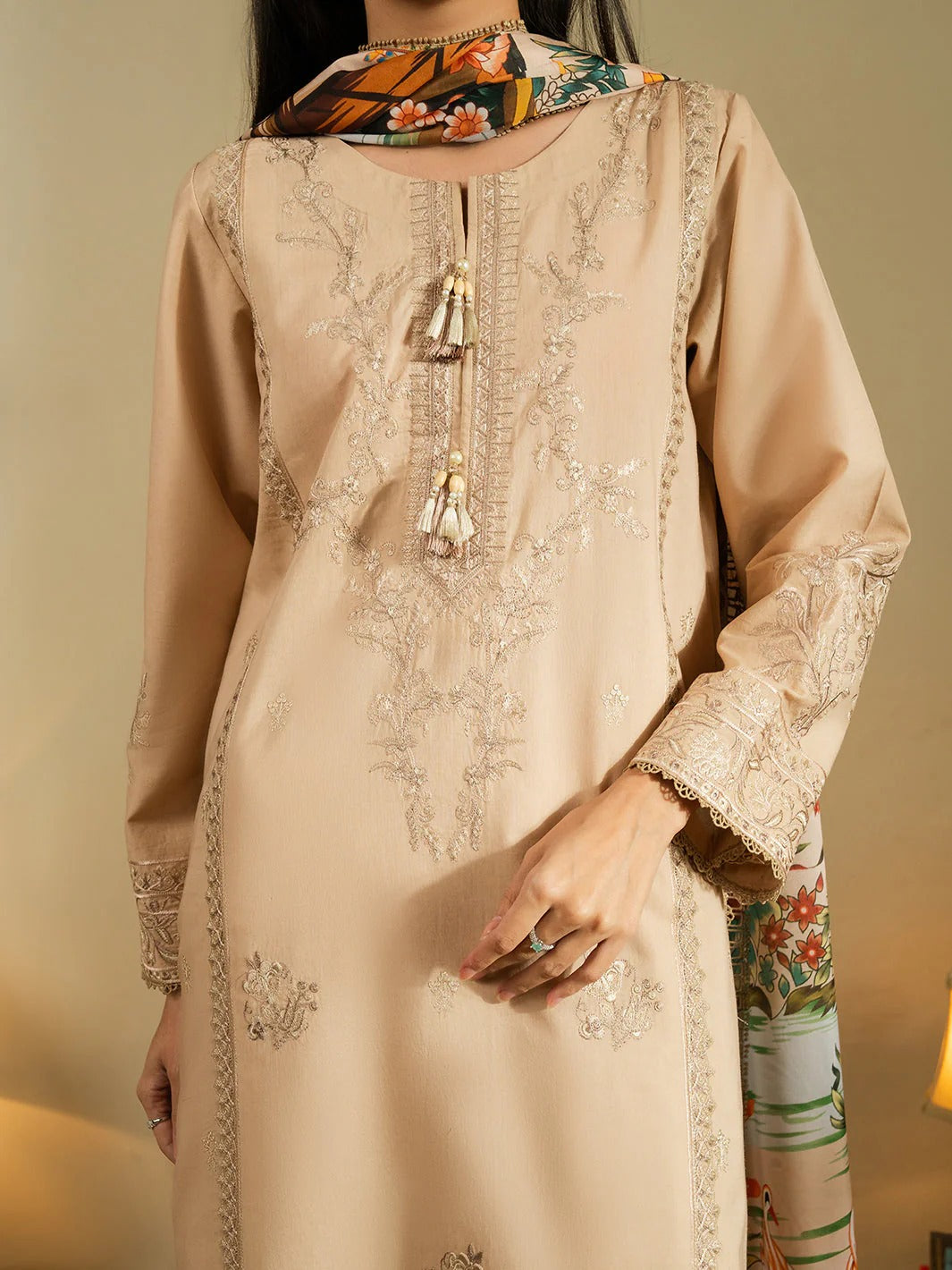A woman wearing a beige embroidered kurta with floral embroidery and tassel details, paired with a multicolor patterned scarf, shown against a warm neutral background.