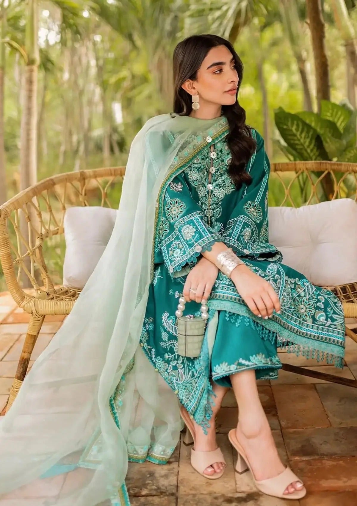 A woman seated on a wicker chair outdoors wearing a teal embroidered traditional outfit with a sheer mint dupatta, holding a small metallic clutch bag.