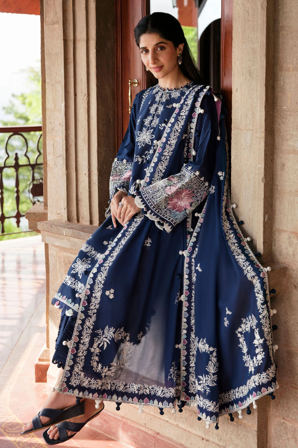 Woman in a blue embroidered traditional outfit standing in a decorative interior setting.