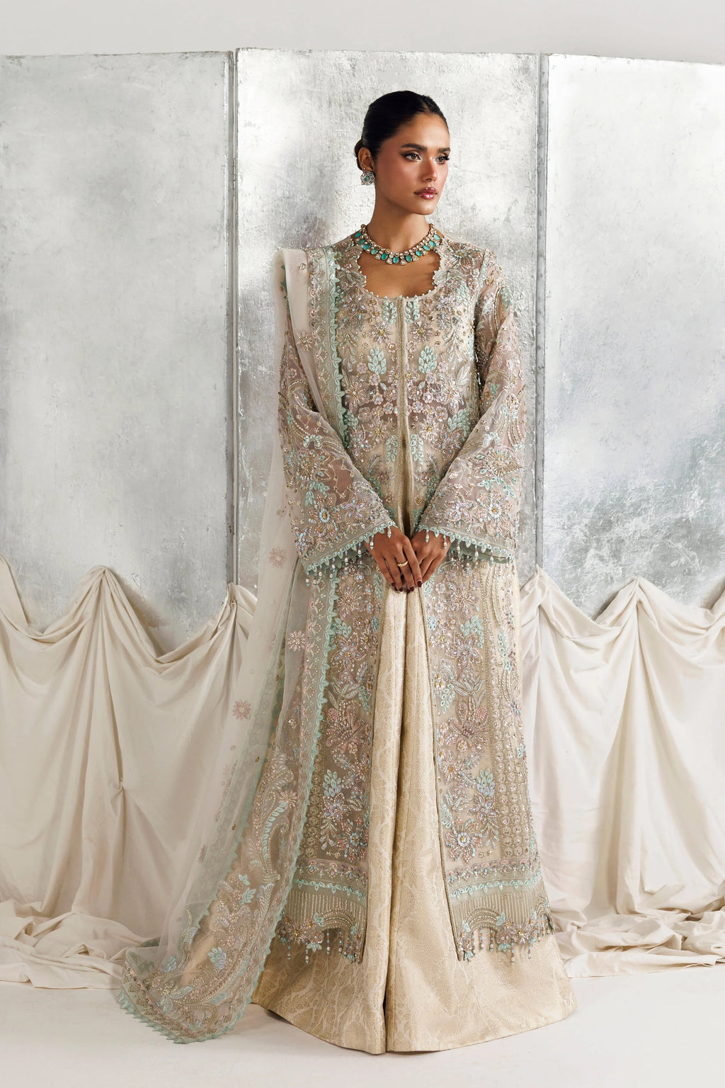 A woman models a traditional South Asian outfit consisting of a beige embroidered kurta, a matching sheer embroidered dupatta, and a turquoise necklace, set against a draped studio backdrop.