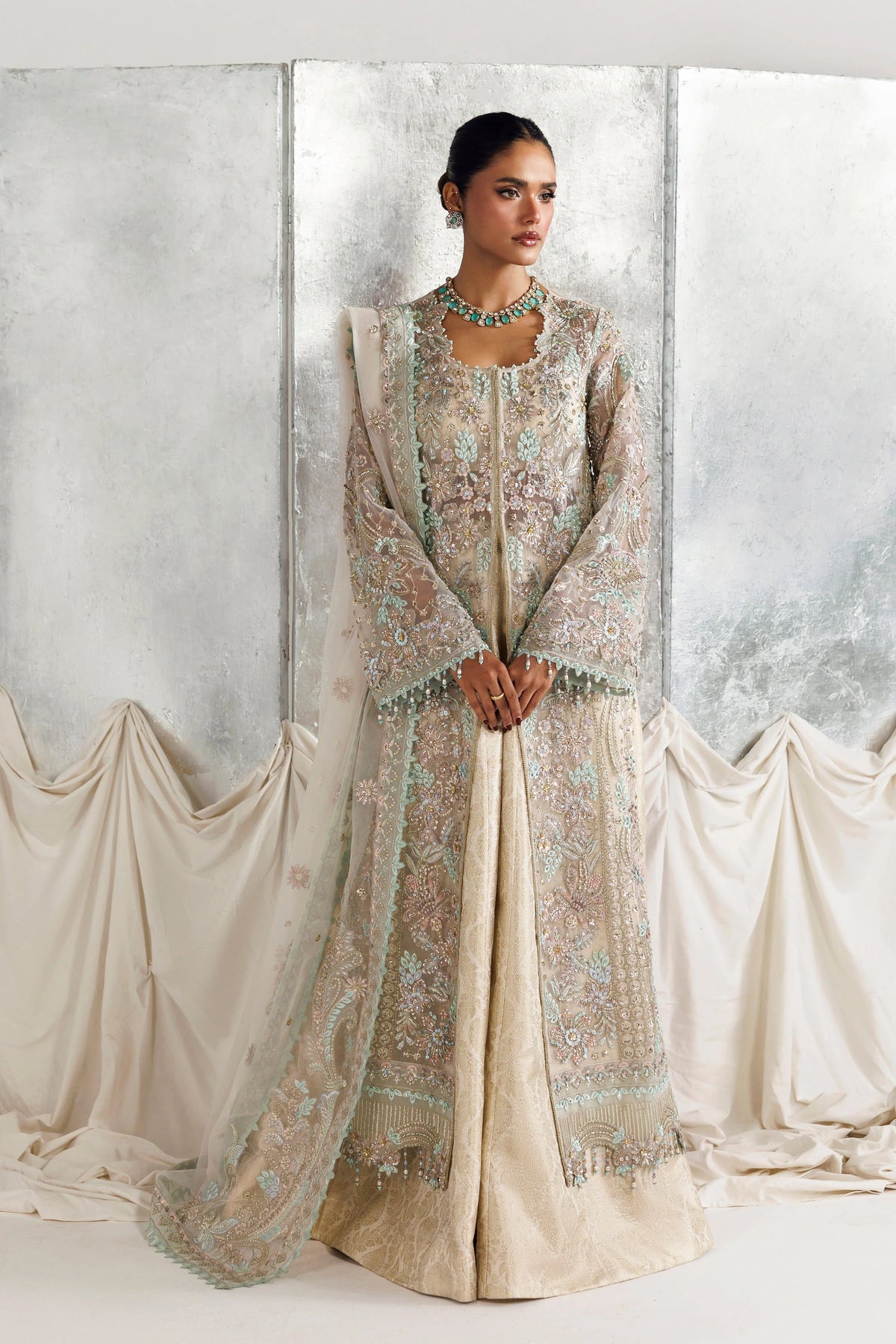 A woman models a traditional South Asian outfit consisting of a beige embroidered kurta, a matching sheer embroidered dupatta, and a turquoise necklace, set against a draped studio backdrop.