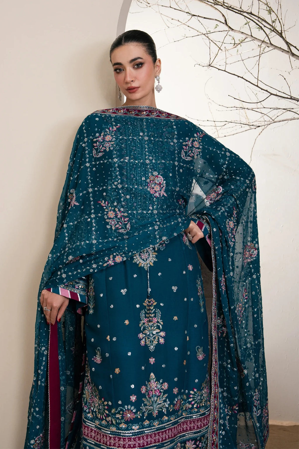 A woman models a traditional teal blue ethnic outfit with intricate floral embroidery and a matching sheer dupatta, set against a neutral indoor background with a decorative branch.