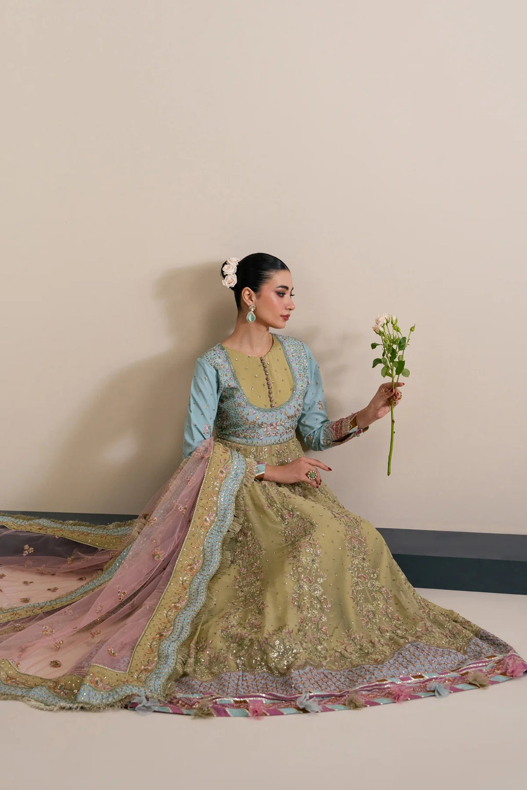 A woman wearing an ornate traditional outfit sits against a plain beige wall, holding a small bouquet of white flowers. The outfit features a light blue and olive green embroidered top with a voluminous pink and green skirt and dupatta.