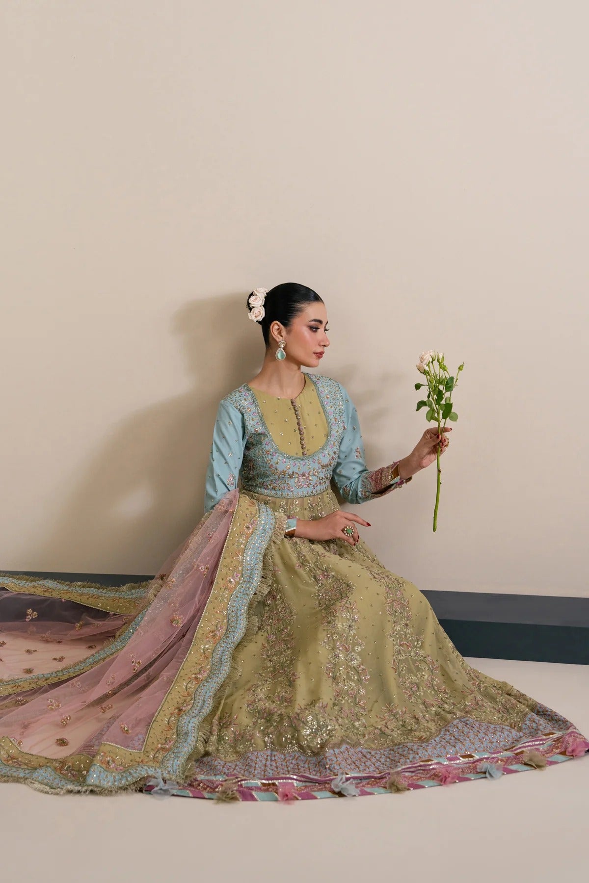A woman wearing an ornate traditional outfit sits against a plain beige wall, holding a small bouquet of white flowers. The outfit features a light blue and olive green embroidered top with a voluminous pink and green skirt and dupatta.