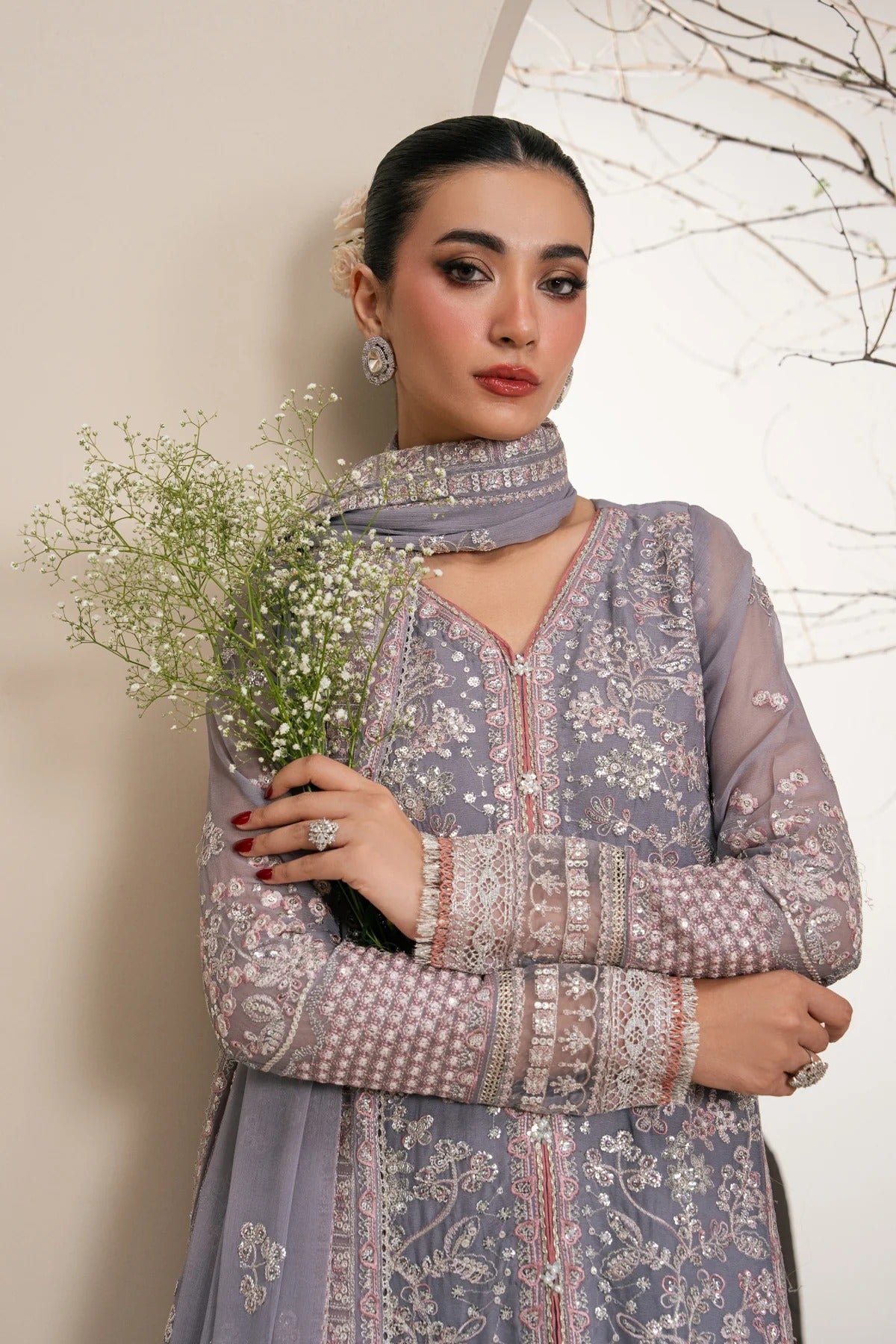A woman models a lavender-gray embroidered traditional outfit with a matching scarf, holding a small bouquet of baby's breath against a light beige wall with delicate branch artwork.