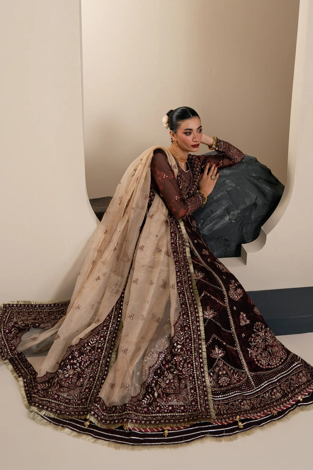 A woman models a traditional Indian outfit consisting of a dark plum velvet anarkali-style dress with intricate gold embroidery, a matching embroidered dupatta, and a striped skirt, set against a minimalist beige studio background.