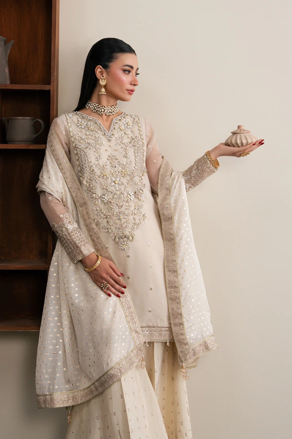 A woman models a traditional cream-colored embroidered ethnic outfit with sheer sleeves and a matching dupatta, standing indoors against a light beige wall with a wooden shelf on the left.