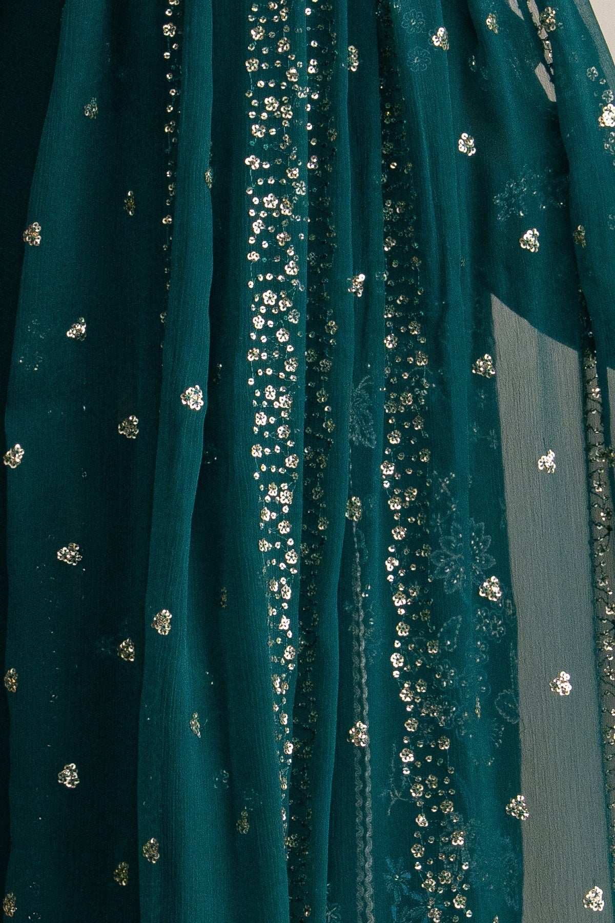 A close-up of a dark teal green sheer fabric, likely a dupatta or saree pallu, decorated with vertical rows of silver floral sequins and scattered silver floral motifs.