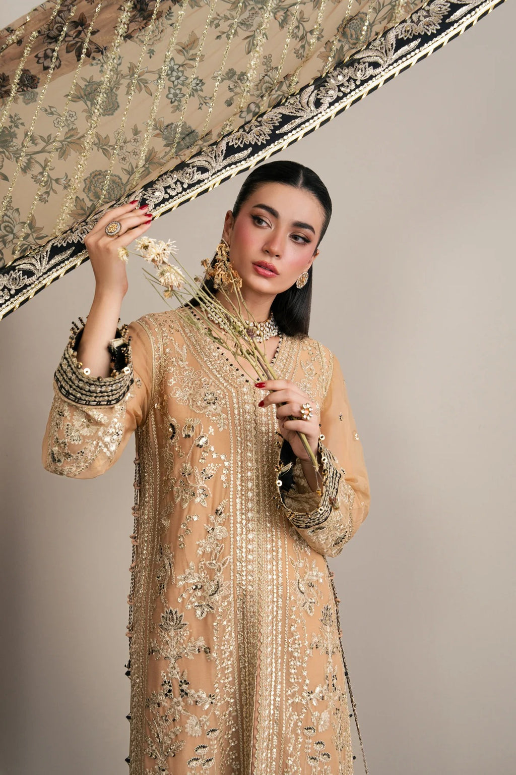 A woman models a beige embellished traditional outfit with intricate gold embroidery, holding a matching patterned dupatta against a plain beige studio background.
