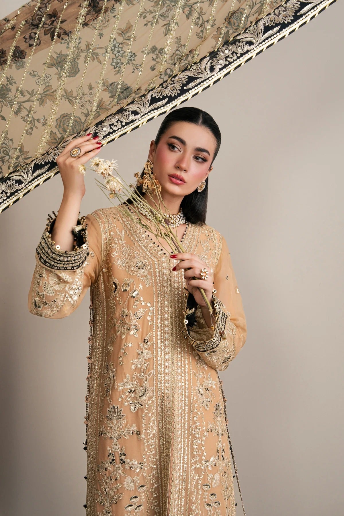 A woman models a beige embellished traditional outfit with intricate gold embroidery, holding a matching patterned dupatta against a plain beige studio background.