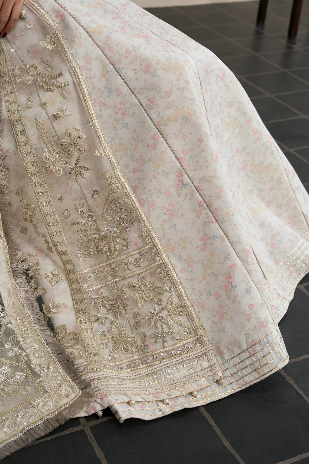 A close-up of a light cream or off-white embroidered fabric, likely a traditional garment or textile, featuring detailed metallic gold floral embroidery and a delicate pastel floral print. The fabric is draped over a dark tiled floor.