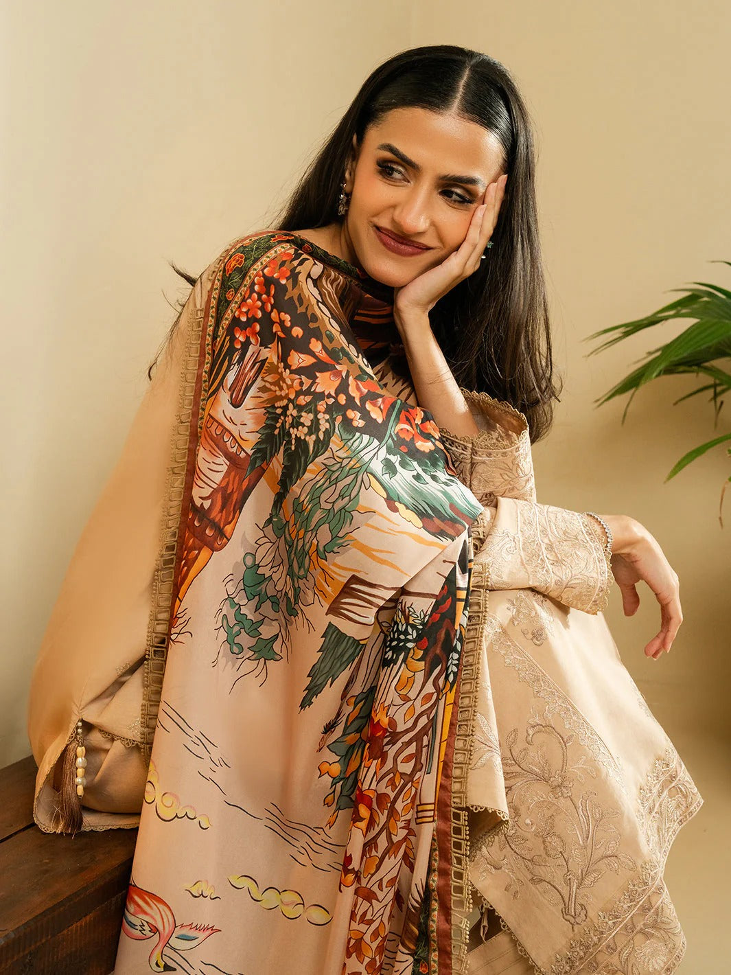A woman wearing a beige printed shawl and matching embroidered kurta, posing indoors against a warm beige wall with a green plant in the background.