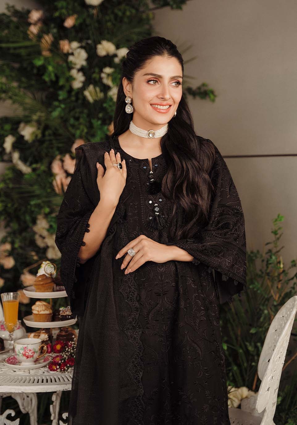 A woman wearing a black embroidered tunic dress with wide sleeves and a choker necklace, standing in a decorated indoor setting with floral arrangements and a small table in the background.