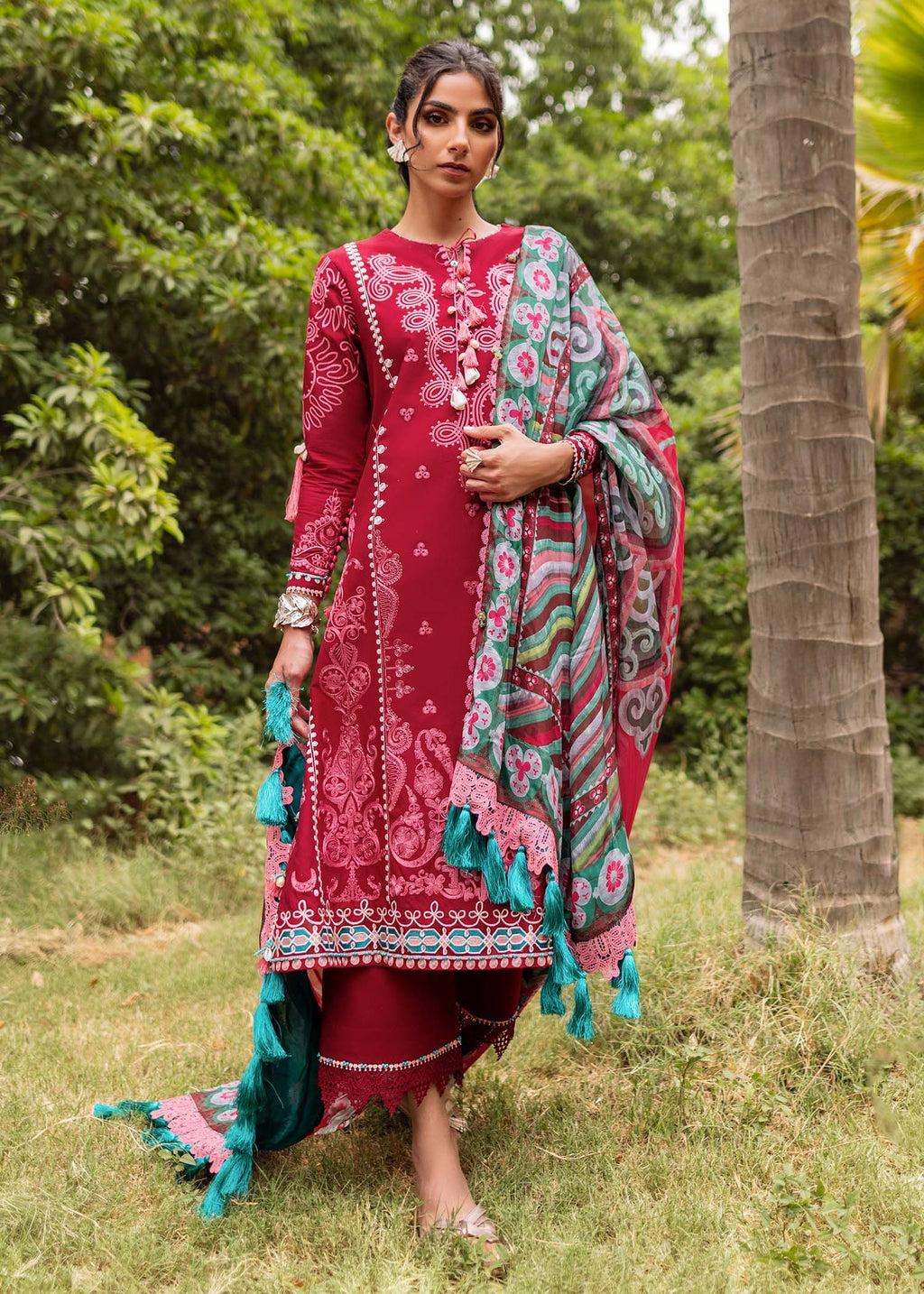 Woman in a red and multicolored traditional outfit standing outdoors with greenery.