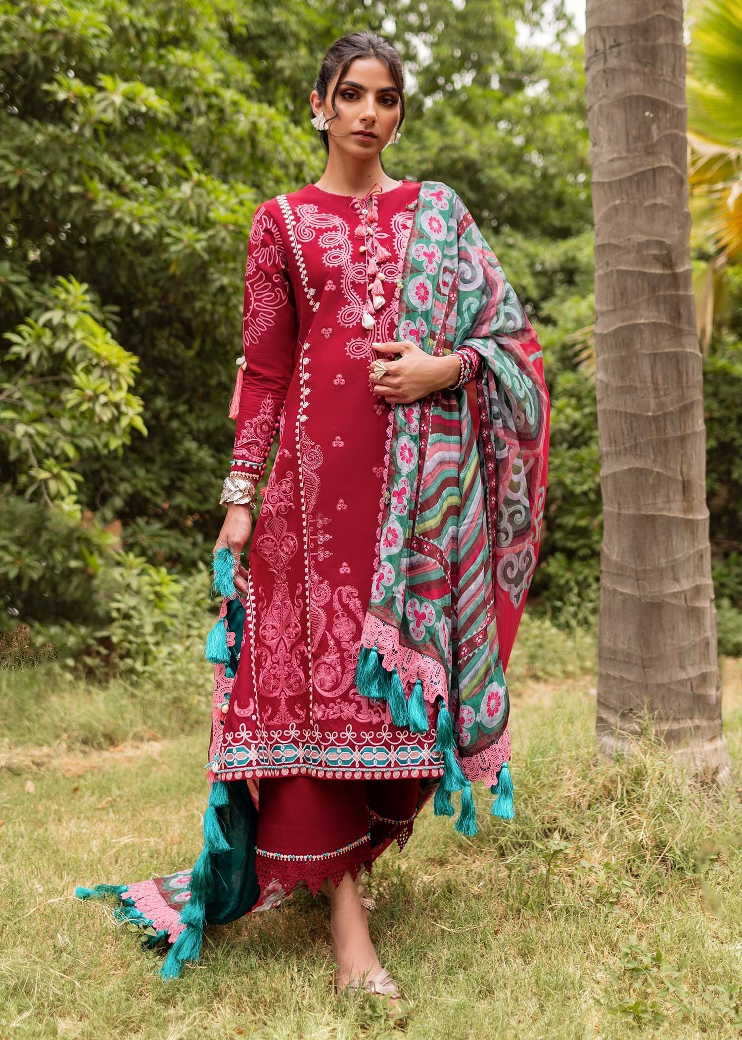 Woman in a red and multicolored traditional outfit standing outdoors with greenery.