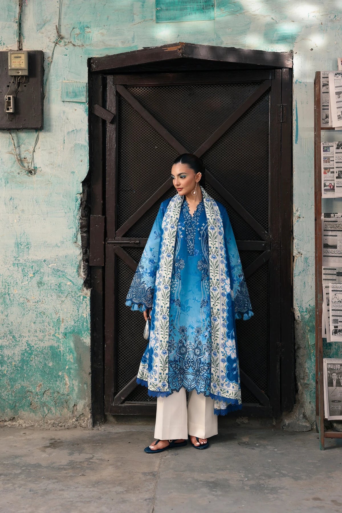 A woman stands in front of a dark wooden door wearing a blue embroidered traditional outfit with white floral patterns, cream wide-leg pants, and black flat sandals, set against a weathered teal wall.