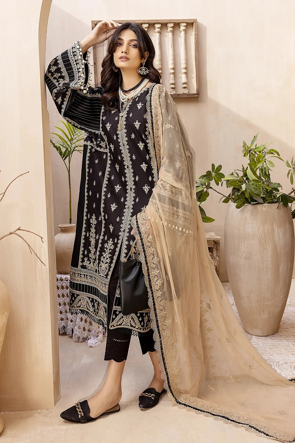 A woman models a black three-piece salwar kameez suit with intricate silver embroidery, paired with a beige sheer net dupatta and black flat shoes, set against a warm beige background with potted plants.