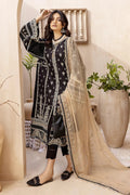 A woman models a black three-piece salwar kameez suit with intricate silver embroidery, paired with a beige sheer net dupatta and black flat shoes, set against a warm beige background with potted plants.