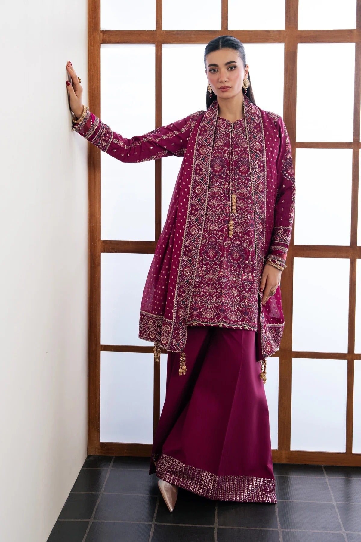 A woman models a traditional South Asian outfit consisting of a deep magenta embroidered kurta and matching flared trousers. She is standing in a corner with a white wall, wooden-framed window, and dark tiled floor.