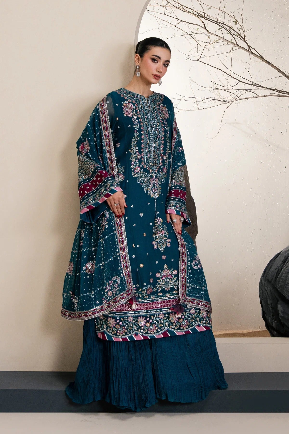 A woman models a traditional South Asian outfit featuring a deep teal blue kurta heavily embroidered with pink, gold, and white floral patterns, paired with matching pleated skirt and a draped dupatta.