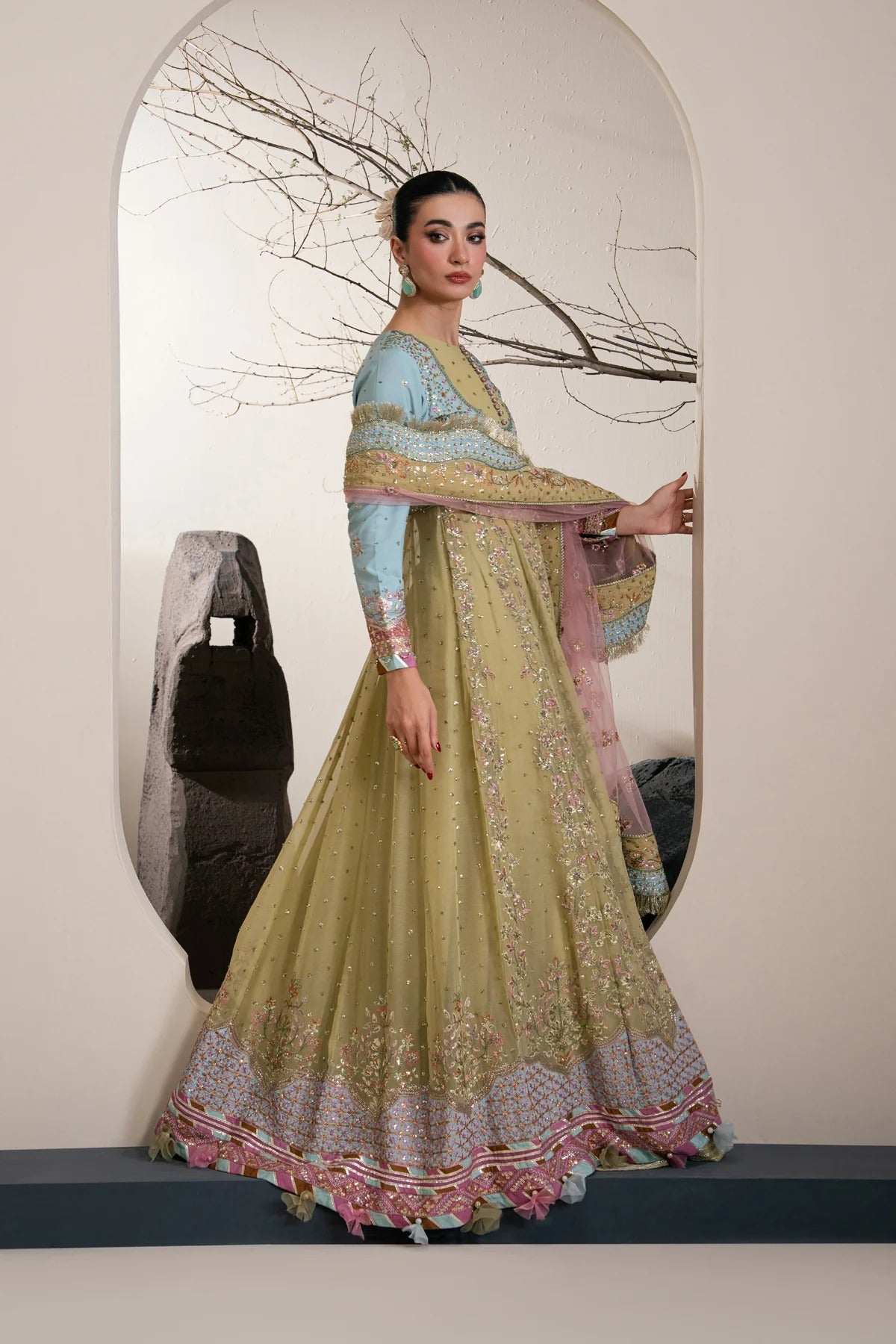 A woman models a traditional Indian anarkali-style dress with a light olive green flared skirt, light blue sleeves, and a pink and gold patterned dupatta, posed in front of a large arched mirror.