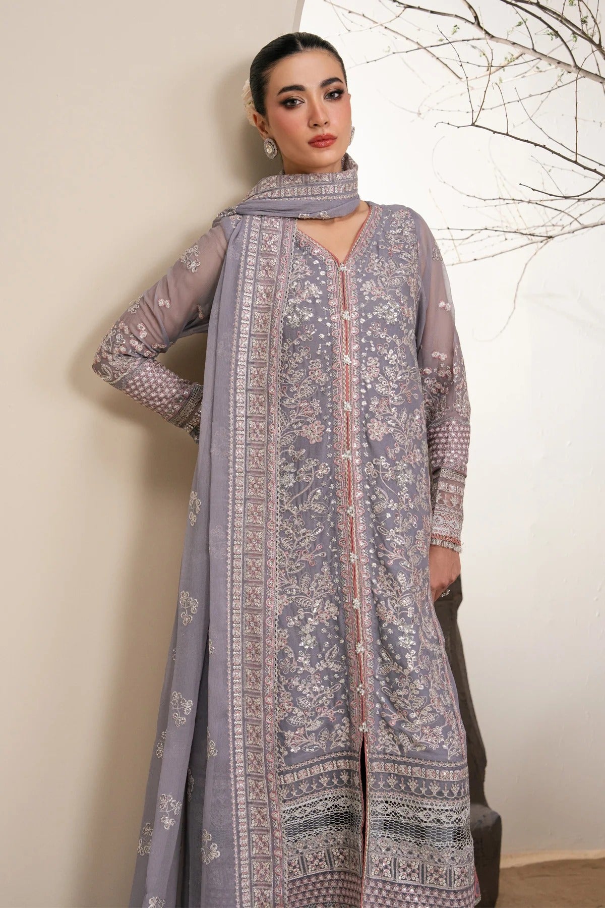 A woman models a traditional three-piece salwar kameez set in a dusty lavender-gray color with intricate silver and white embroidery. The set includes a long kurta, a matching dupatta, and a coordinating long overcoat draped over her arm.