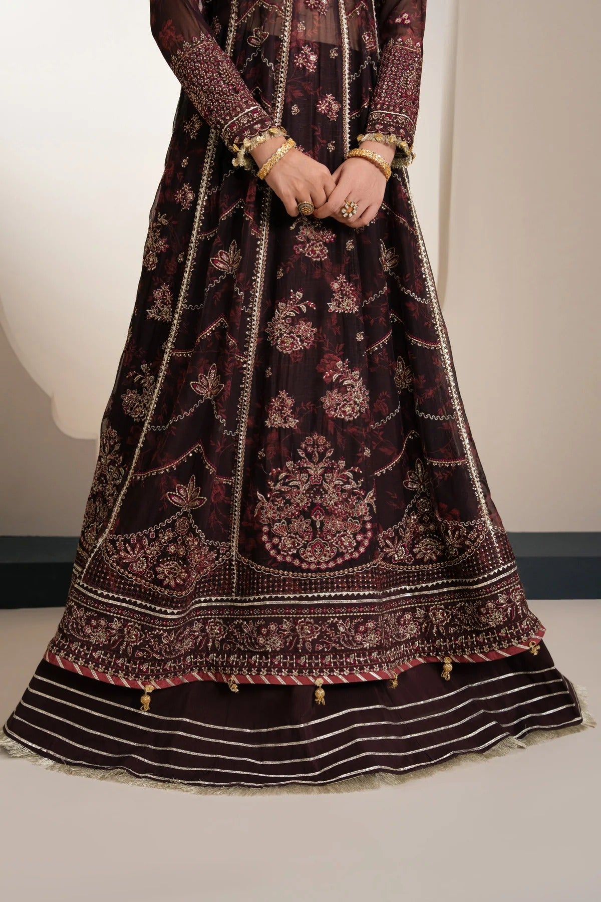 A dark plum embroidered Anarkali gown with intricate gold and pink floral patterns, paired with a matching striped skirt, shown on a model.