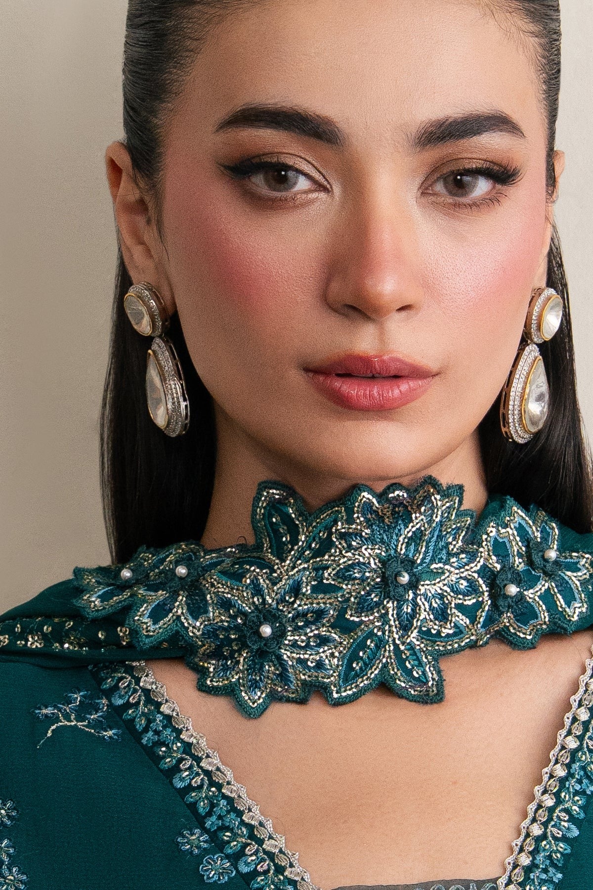 A close-up portrait of a woman wearing a dark teal embroidered choker necklace with silver beadwork, matching teal embroidered clothing, and large silver teardrop earrings against a soft beige background.