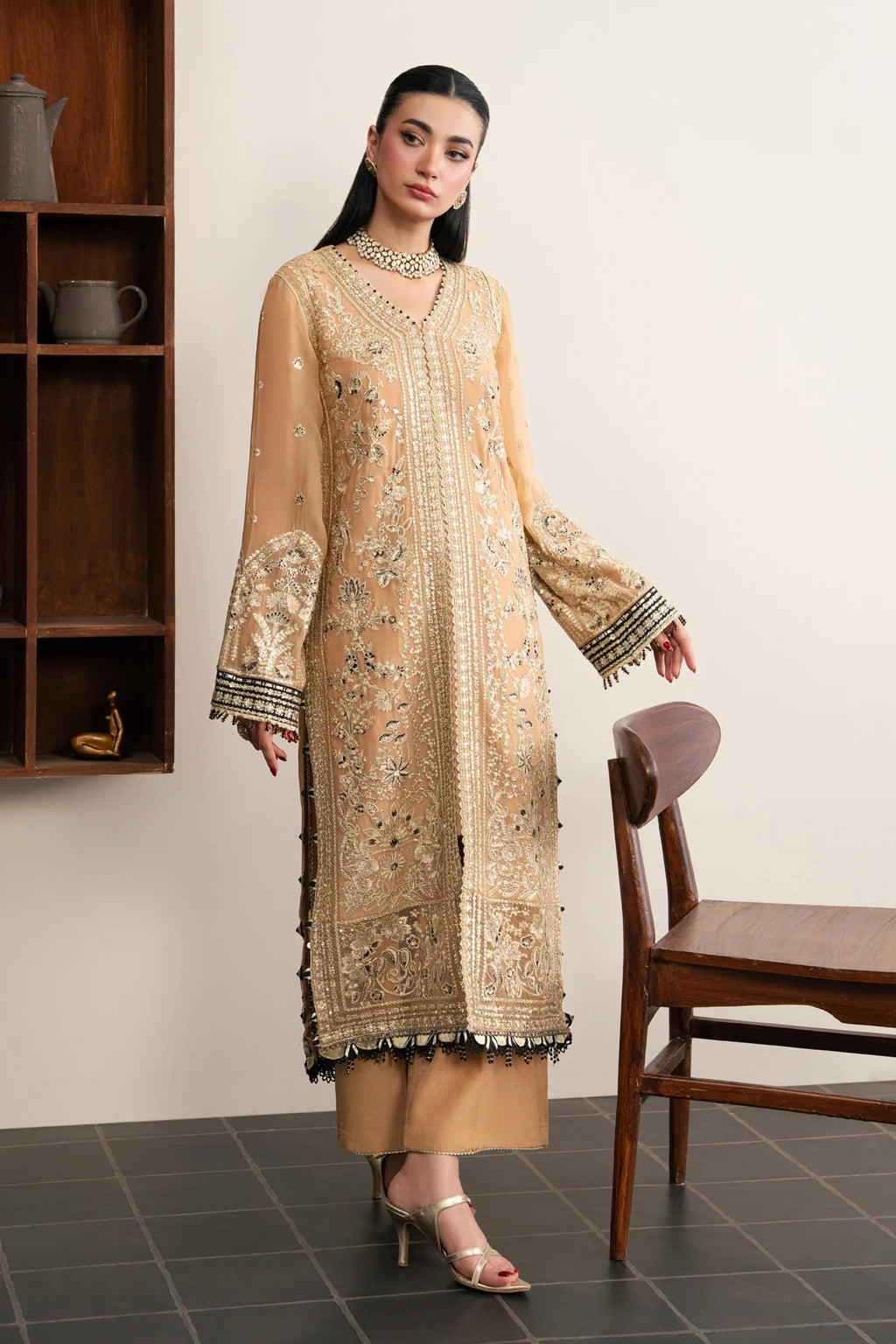 A woman models a beige embroidered traditional kurta set with matching pants and heels, standing in a room with a wooden shelf and chair.