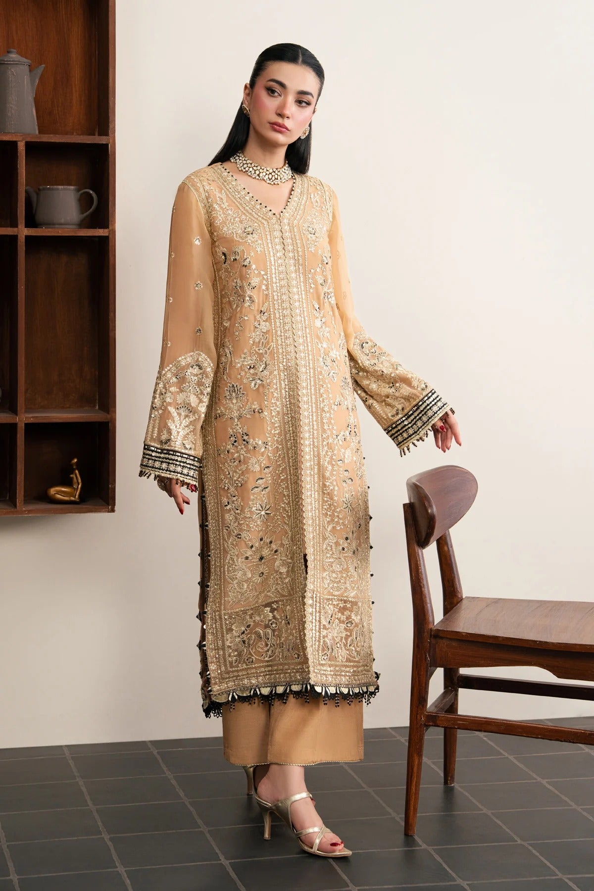 A woman models a beige embroidered traditional kurta set with matching pants and heels, standing in a room with a wooden shelf and chair.