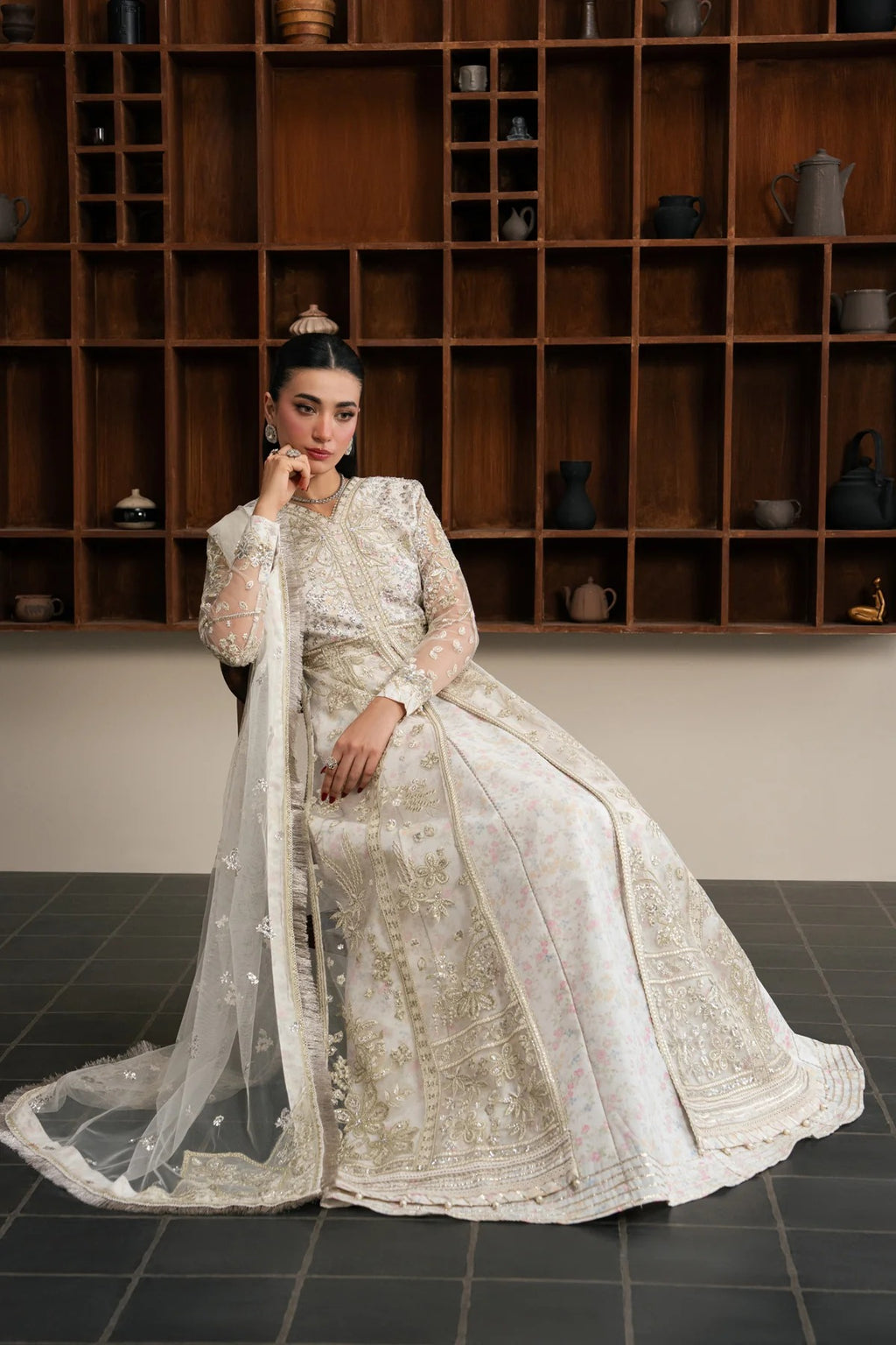 A woman models a traditional white and gold embroidered Anarkali gown with a matching sheer dupatta, posed in front of a dark wooden shelving unit.