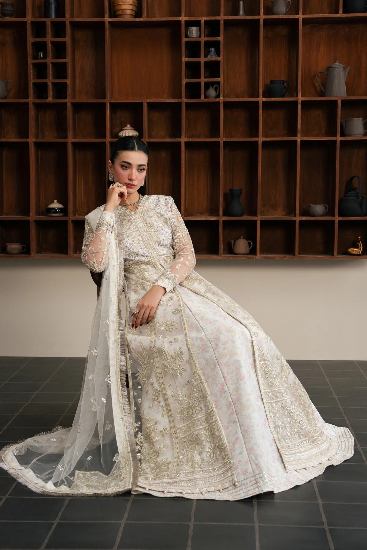 A woman models a traditional white and gold embroidered Anarkali gown with a matching sheer dupatta, posed in front of a dark wooden shelving unit.