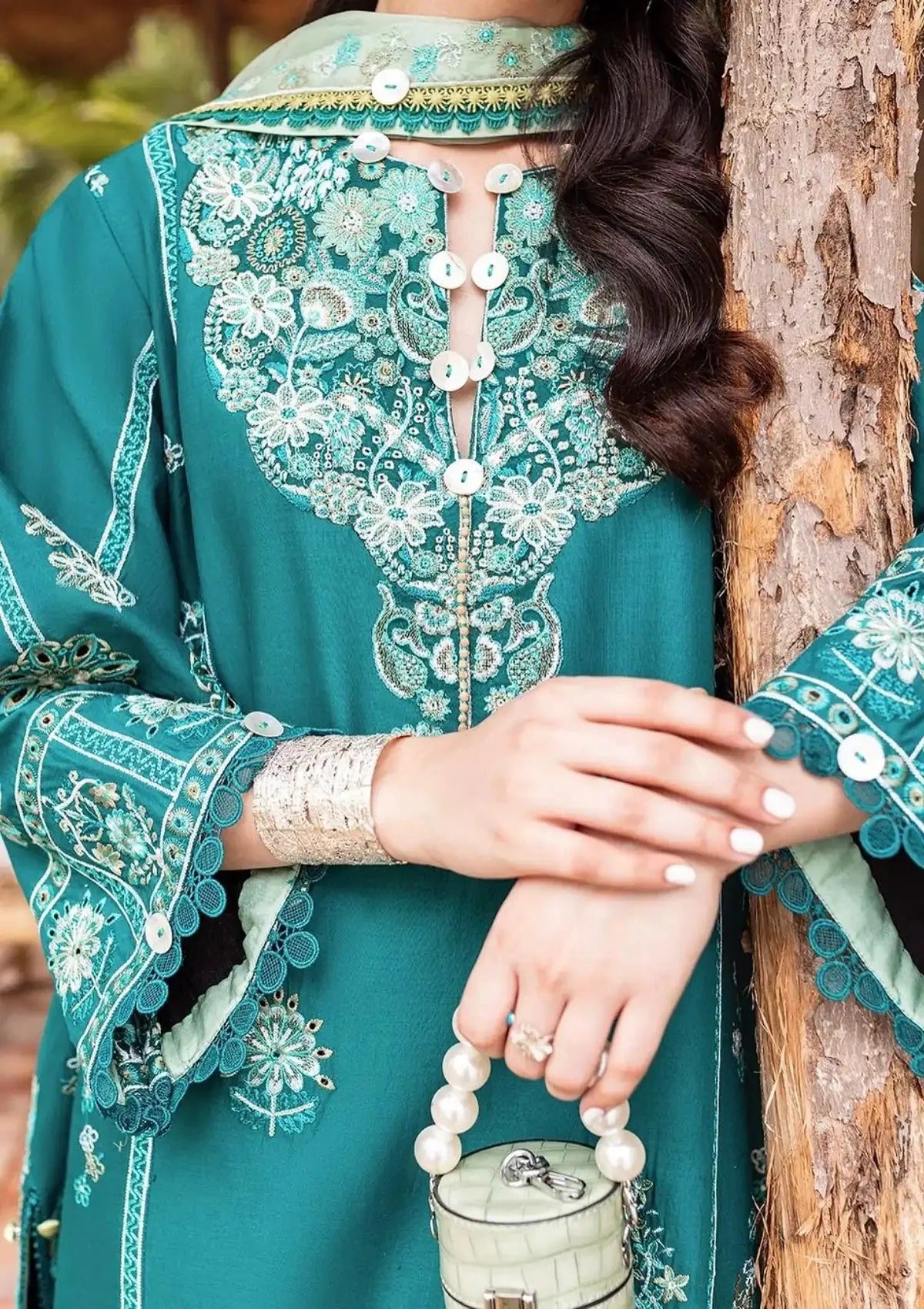 A close-up of a woman wearing a teal green embroidered traditional outfit with intricate white floral embroidery, holding a small pearl handbag next to a tree trunk.