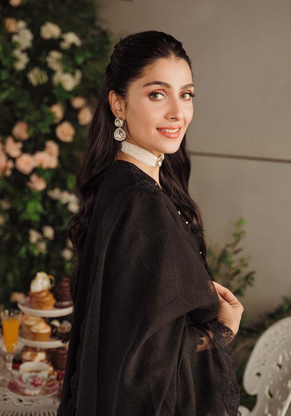 Woman in a black outfit with lace details standing in front of floral decorations and pastries.