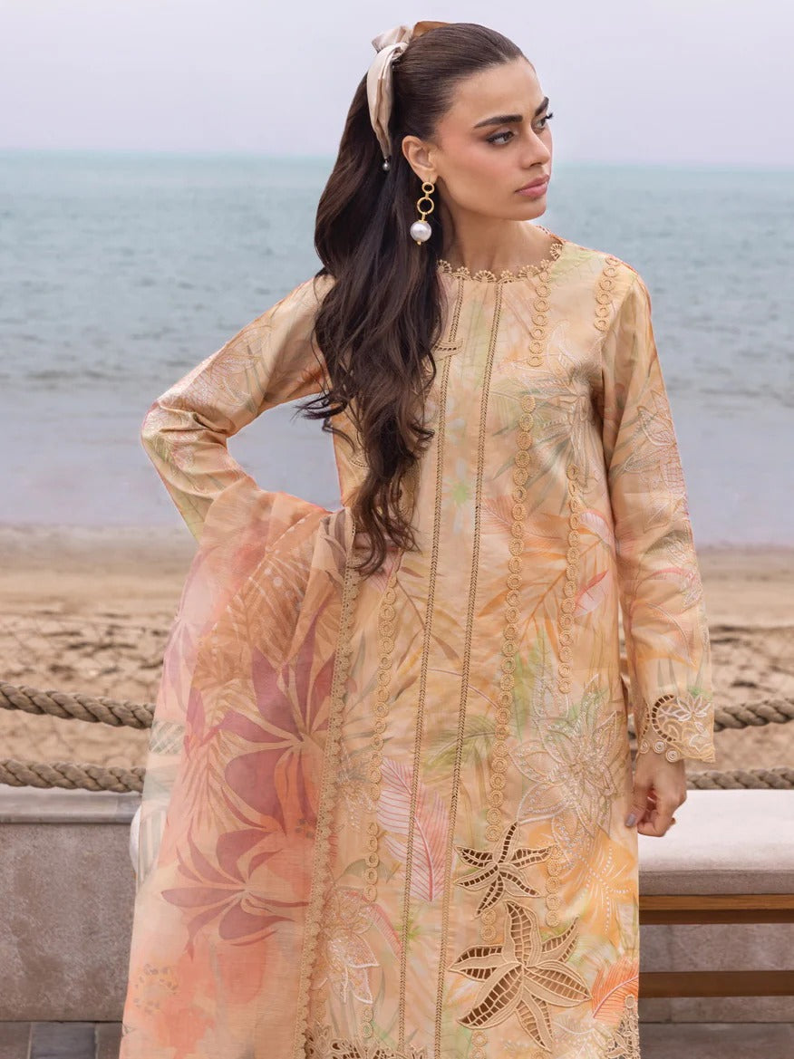 A woman wearing a beige printed traditional outfit with floral patterns and gold embroidery, standing outdoors near the beach with the ocean in the background.