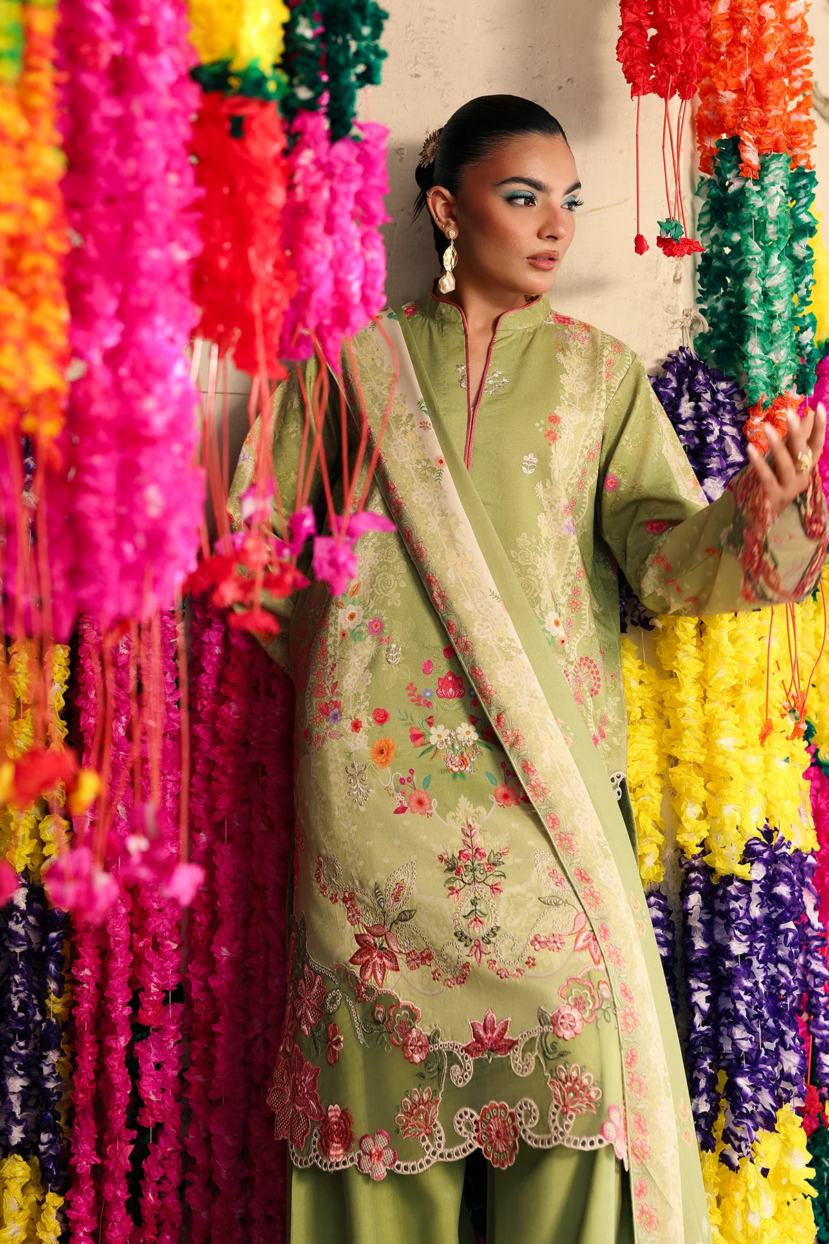 A woman models a light olive green embroidered traditional outfit with a matching dupatta, set against a colorful background of hanging flower garlands.