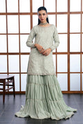 A woman models a traditional three-piece salwar kameez suit in a sage green color. The set includes an embroidered long-sleeve kameez top, matching tiered palazzo pants, and a coordinating belt, set against a wooden lattice screen background.