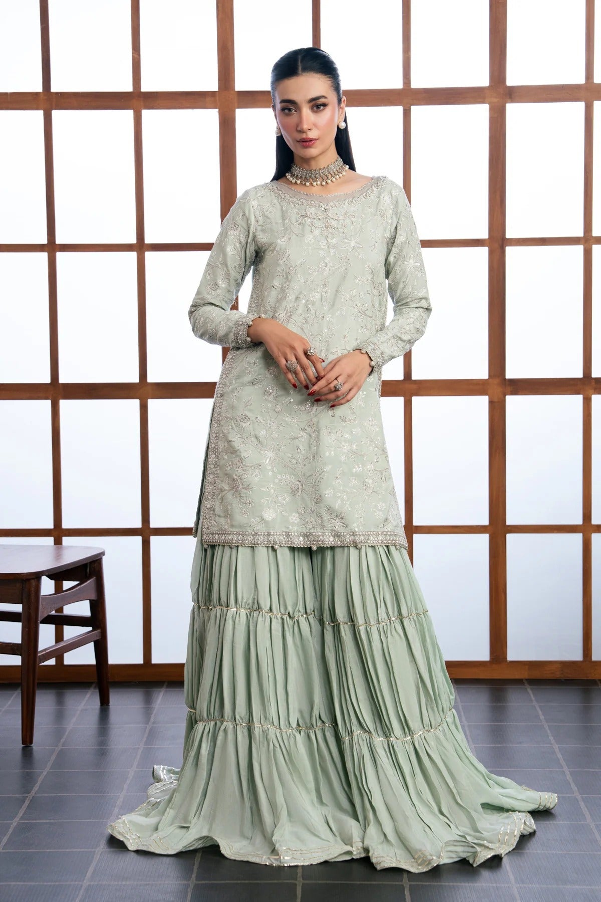 A woman models a traditional three-piece salwar kameez suit in a sage green color. The set includes an embroidered long-sleeve kameez top, matching tiered palazzo pants, and a coordinating belt, set against a wooden lattice screen background.