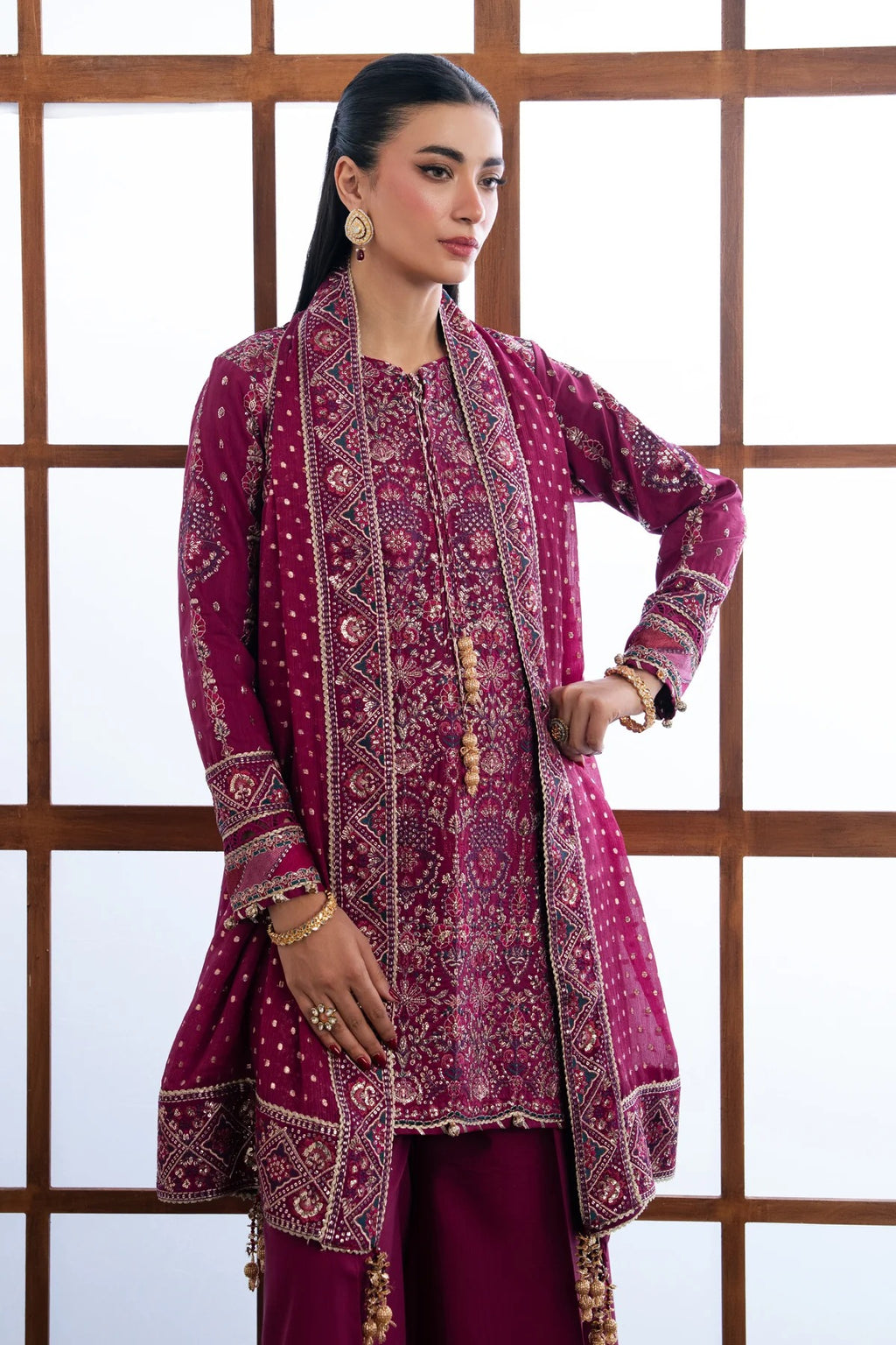 A woman modeling a traditional South Asian outfit featuring a deep magenta embroidered kurta with gold and silver mirror work, paired with matching trousers, set against a white background with a wooden grid screen.