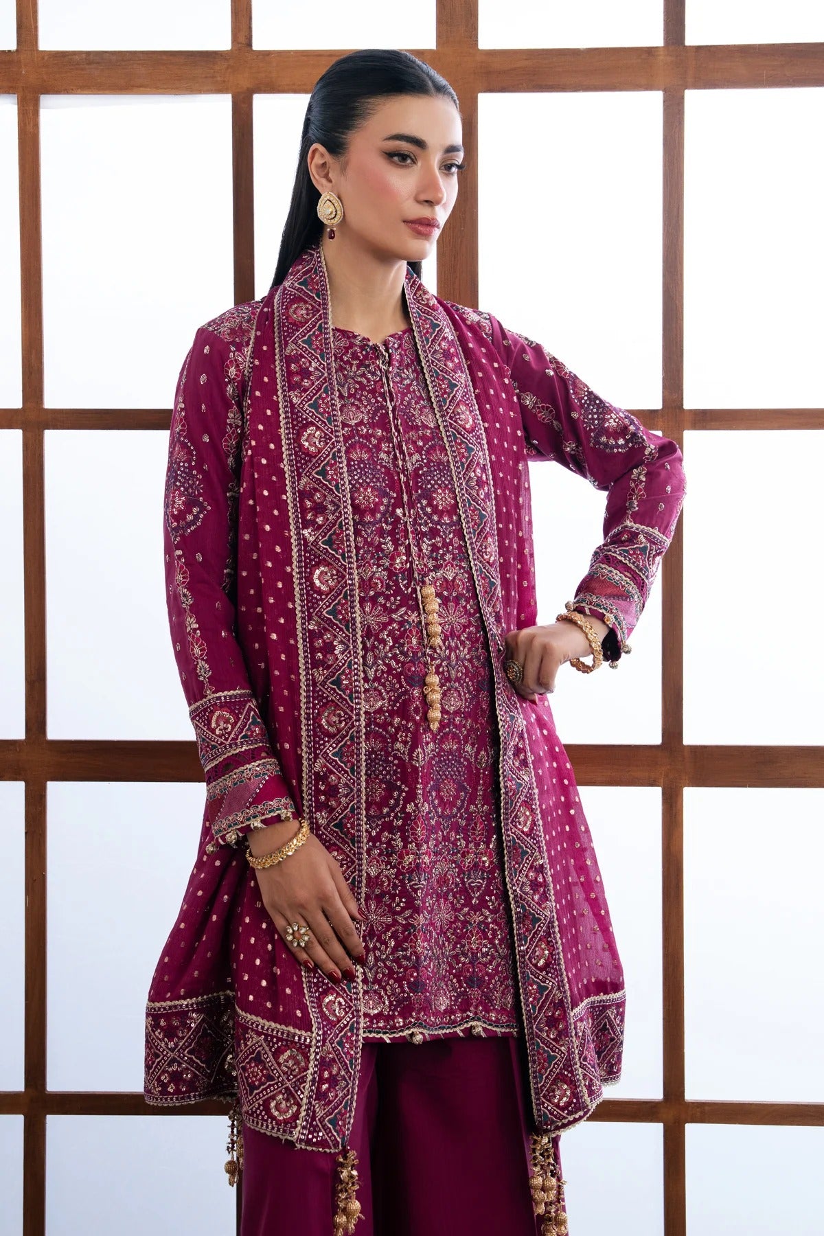 A woman modeling a traditional South Asian outfit featuring a deep magenta embroidered kurta with gold and silver mirror work, paired with matching trousers, set against a white background with a wooden grid screen.