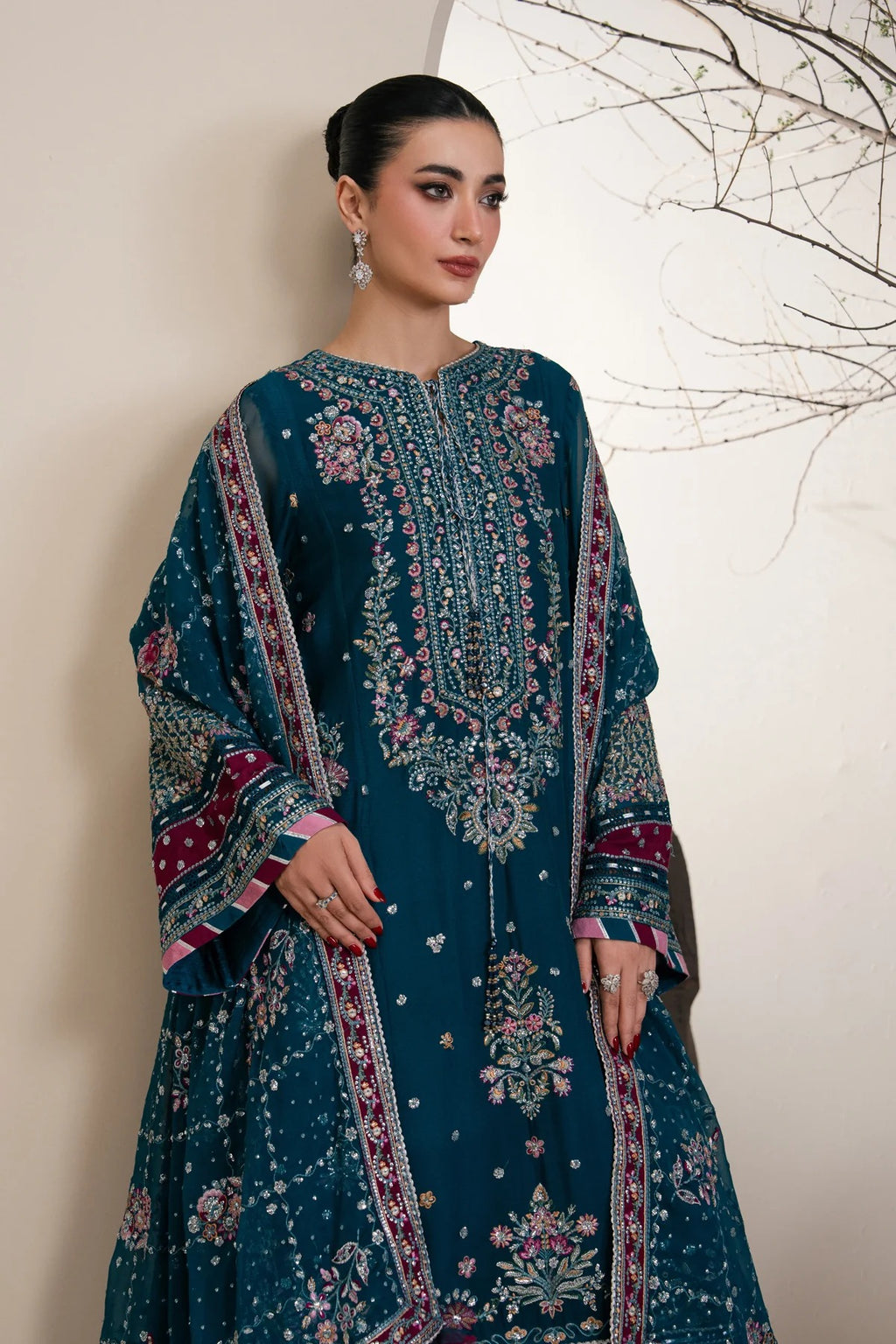 A woman models a dark teal blue traditional embroidered kurta set with intricate floral patterns and colorful borders, set against a neutral beige wall with minimalist branch decor.