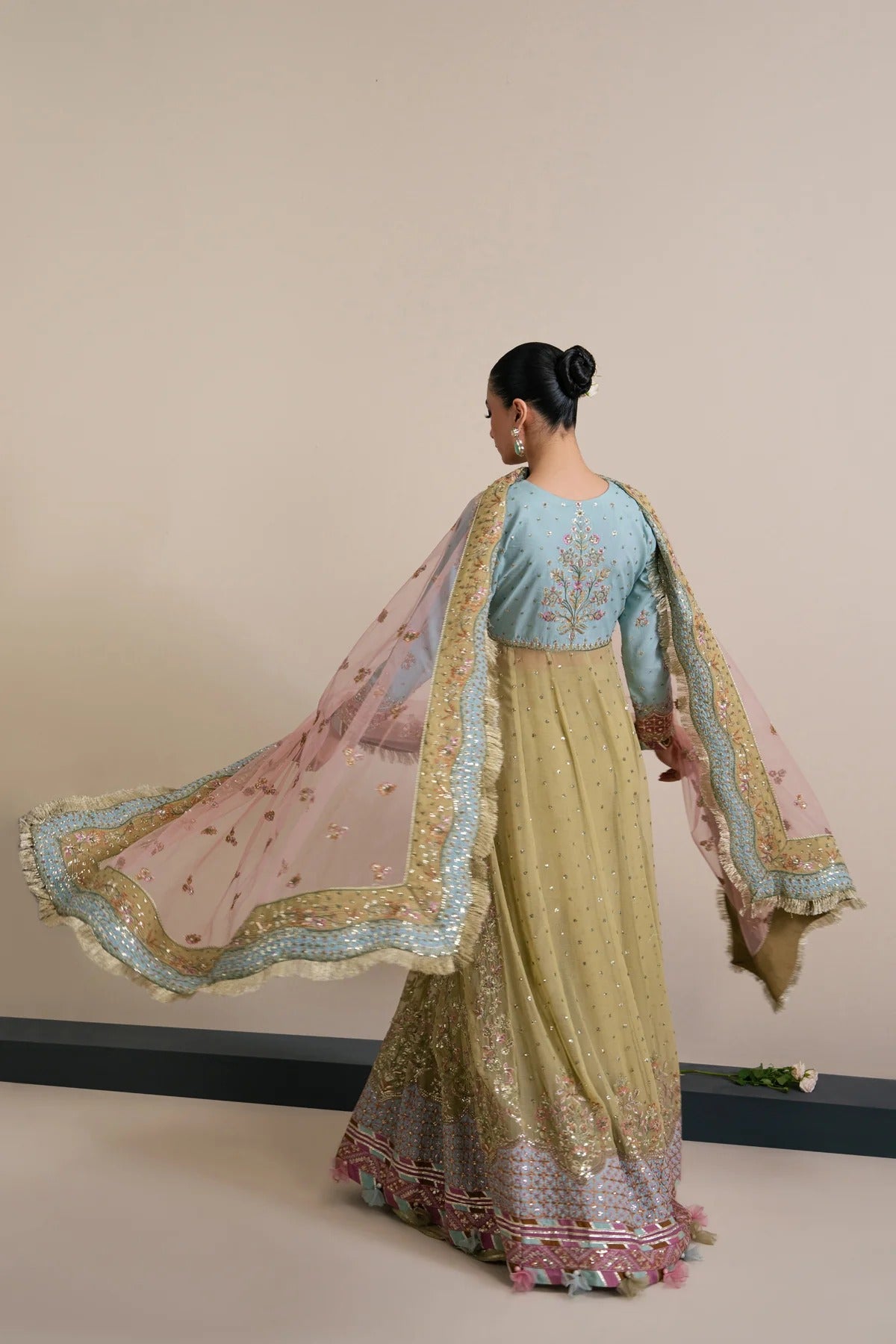 A woman wearing an ornate traditional Indian outfit with a light blue embroidered blouse, a long olive-green skirt with heavy gold embroidery and sequins, and a sheer pink dupatta with gold borders, captured from behind against a plain beige wall.