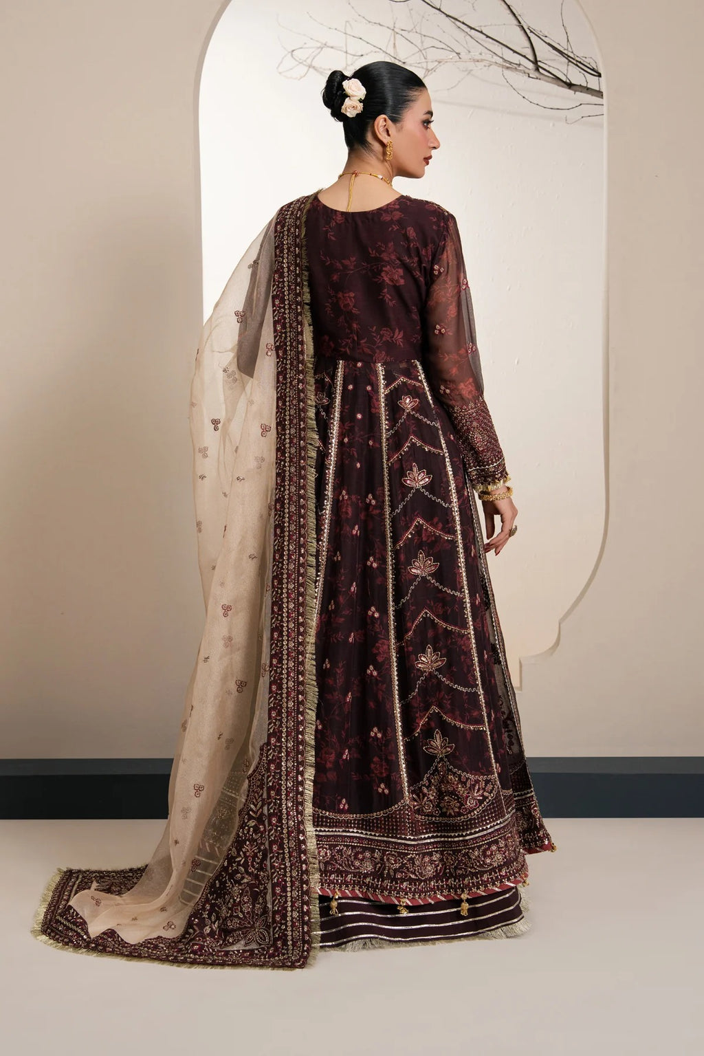 A woman modeling a traditional dark maroon anarkali suit with gold embroidery and a beige printed dupatta, shown from the back in a studio setting.